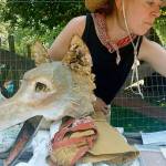 Michelle Lassaline, Mary Olson Farms artist in residence, and Coyote, one of 11 paper-maché masks she has made and dons when she paints for the public, enjoy a cool moment in the welcome shade of an apple tree on her final day at the rebuilt farm last Sunday. ROBERT WHALE, Auburn Reporter